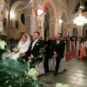 fotografia ślubna przedstawia państwa młodych siedząch w kościele, w tle goście ślubni